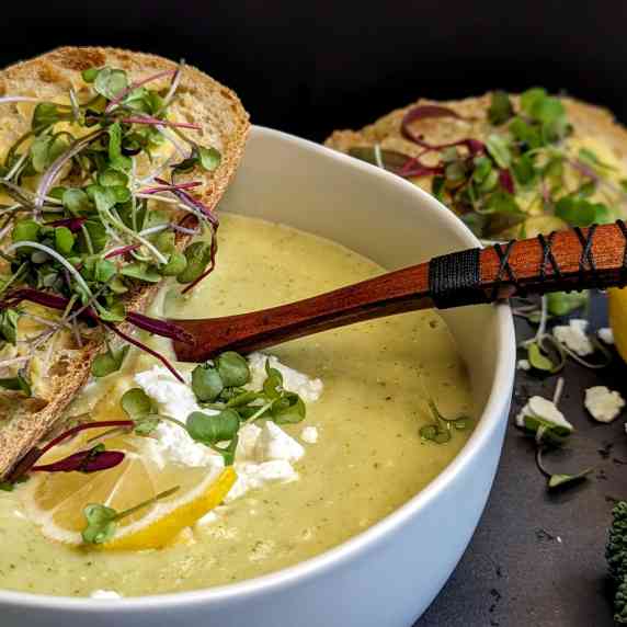 white bowl with green soup, topped with a crusty bread with butter and sprouts, in the background, s