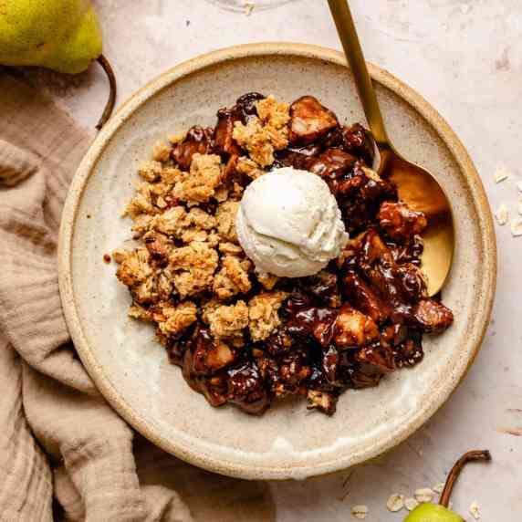 A bowl of pear and chocolate crumble with ice cream on top and a spoon, with pears and oats around.