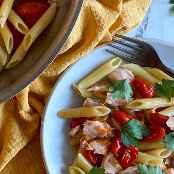 Image shows a plate of penne pasta with burst cherry tomatoes, flaked salmon, and parsley.