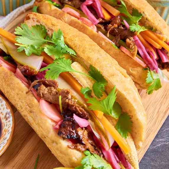 Two Pork Banh Mi buns on a board, loaded with pulled pork, pickled vegetables, and cilantro. 