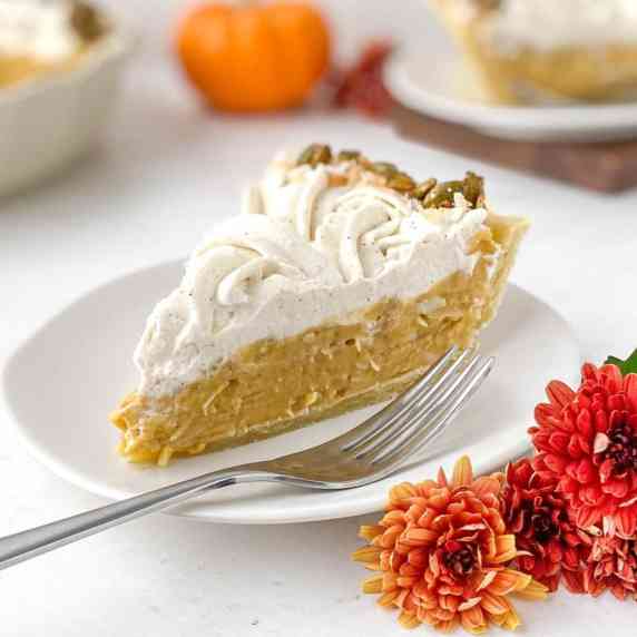 A slice of pumpkin coconut cream pie on a white plate with red and orange mums nearby