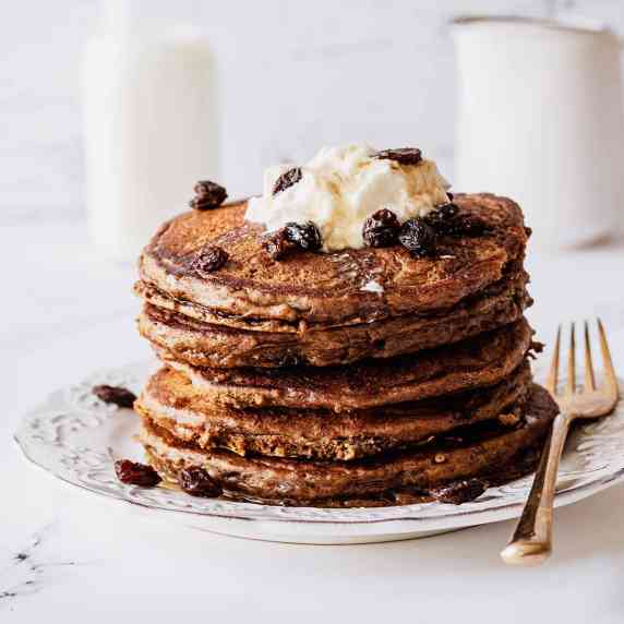 Stack of raisin pancakes topped with whipped cream and raisins on a white plate with a gold fork