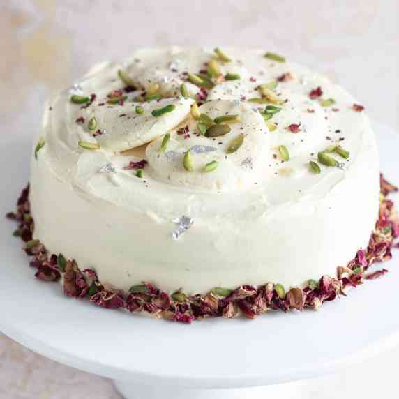 a fresh cream cake is placed on a white cake stand and decorated with rose petals and nuts