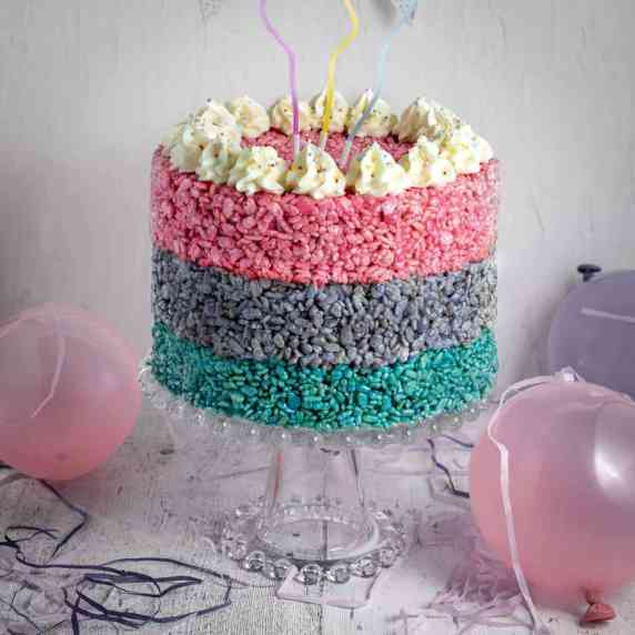 A three layered Rice Krispie cake on a cake stand, surrounded by balloons.