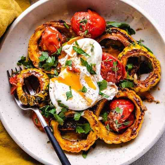 Roasted delicata squash and cherry tomatoes, poached egg, parsley and oil on white plate with fork
