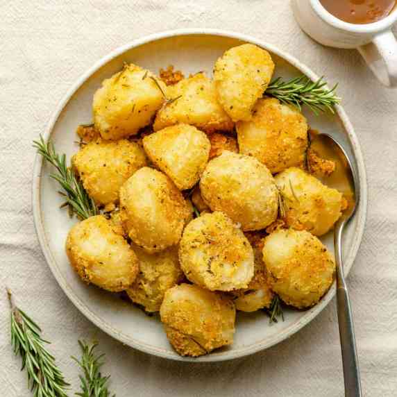 A bowl of crispy rosemary roasted potatoes with a spoon and a jug of gravy on the side.