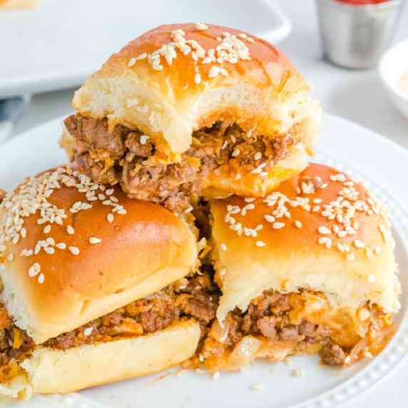 Sloppy Joe sliders stacked on a plate with sesame seeds and the top sandwich is missing a bite.