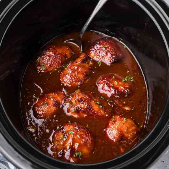 Square view of BBQ chicken thighs in BBQ sauce in a slow cooker with a spoon resting in the sauce.