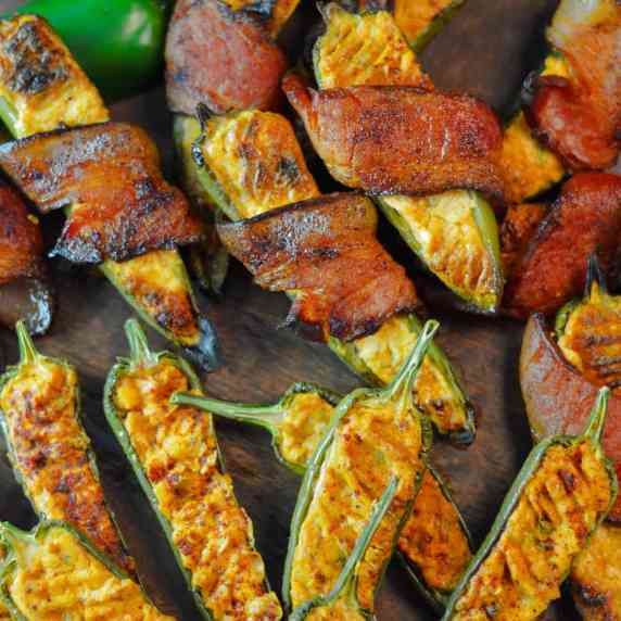 smoked jalapeno poppers on a wooden board, plain and bacon-wrapped with a raw jalapeno in upper left