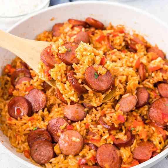 Square view of smoked sausage and rice in a pan with a wooden spoon scooping a spoonful.