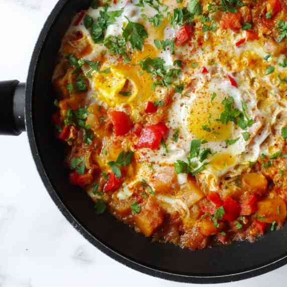 A frying pan sits filled with colorful veggie pisto and is topped with some fried eggs and parsley
