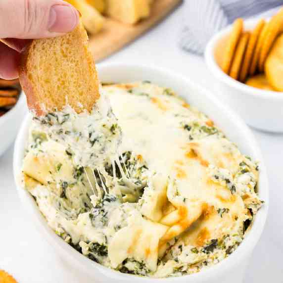 A person's hand dipping bread into a bowl of spinach dip covered in melted cheese.