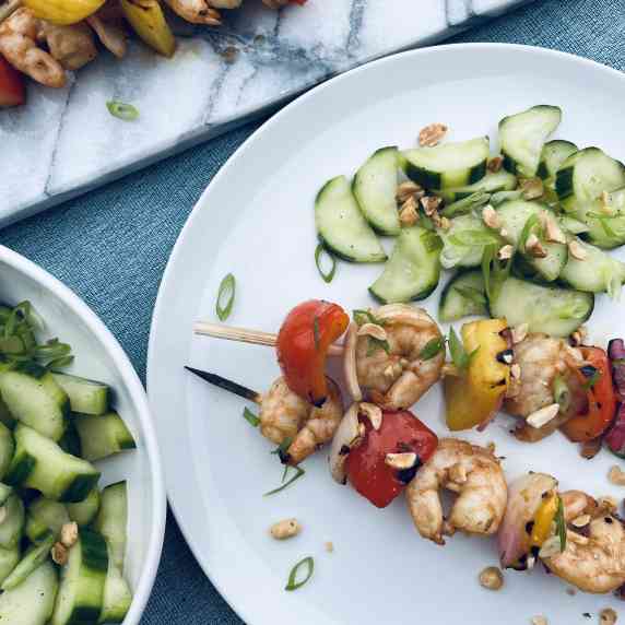 Grilled shrimp and vegetable skewers with marinated cucumbers served on a white plate.
