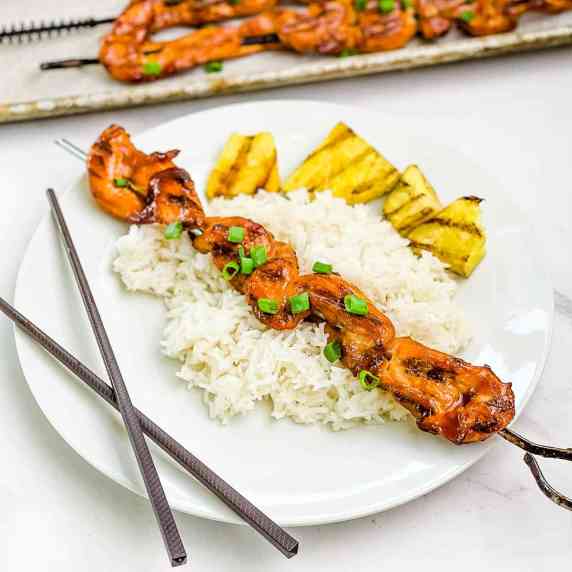 A closeup picture of grilled teriyaki chicken on a stick placed on a plate over white rice.