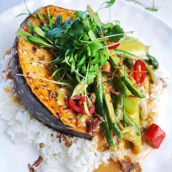Bed of rice on a white plate topped with baked pumpkin, beans, chilli, coriander and red curry sauce