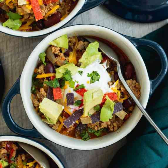 Bowl of turkey chili topped with avocado, cheddar cheese, tortilla strips, and sour cream.