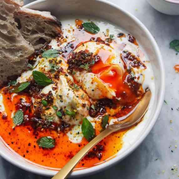 Poached eggs on top of yogurt in a white bowl with chilli oil and a piece of bread.