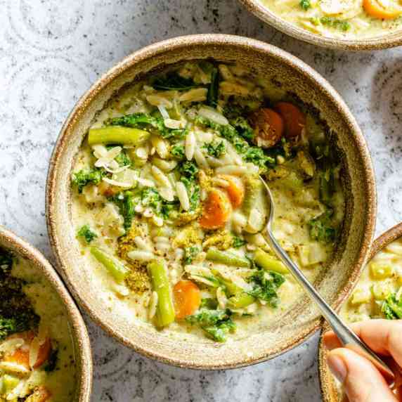 A bowl of vegetable orzo soup with a hand holding a spoon in it. 