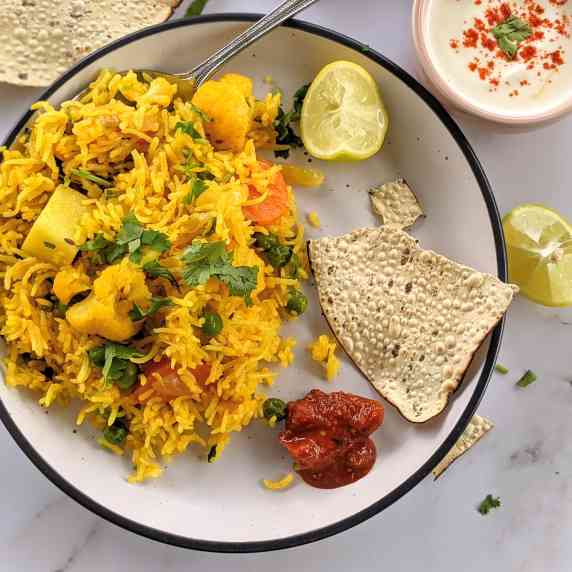 Mixed vegetable rice, papad, pickle, lemon served on a white plate with a bowl of spiced yoghurt.