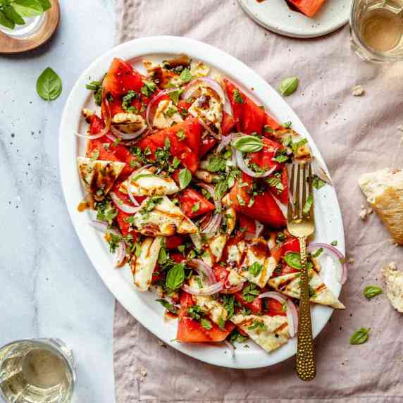 A platter with watermelon, basil, halloumi and herbs with a gold fork on a napkin.