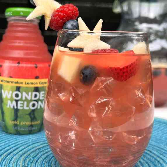 Drink w/ blueberry, apple and strawberry garnishes on a blue placemat w/ Wonder Melon bottle behind