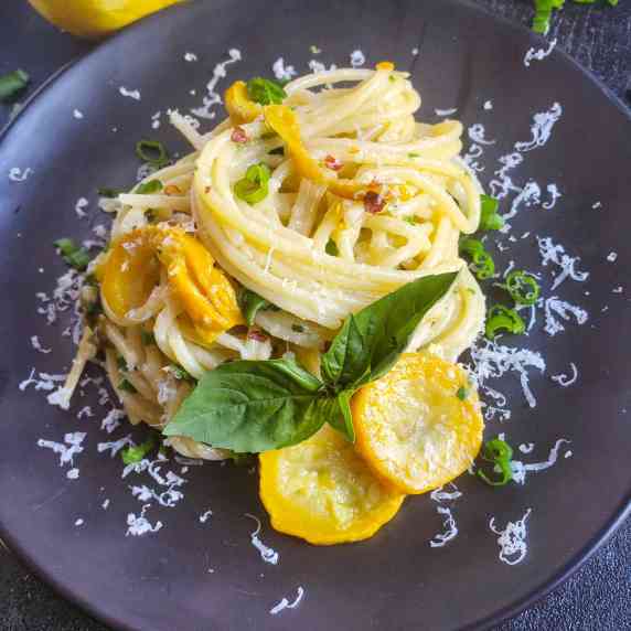 Spaghetti and yellow squash on a black plate with white cheese shredded.