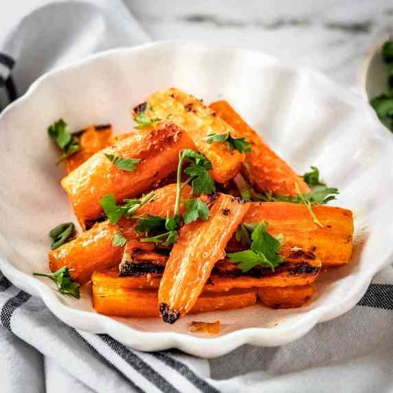 Side shot of roasted carrots garnished with herbs on a white plate on a grey cloth.