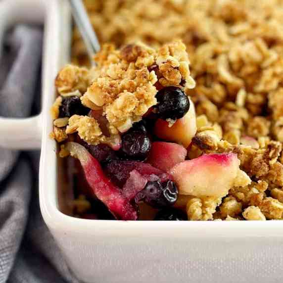 cooked slices of apple and blueberries with oat crumble on top in a white baking dish with a spoon.