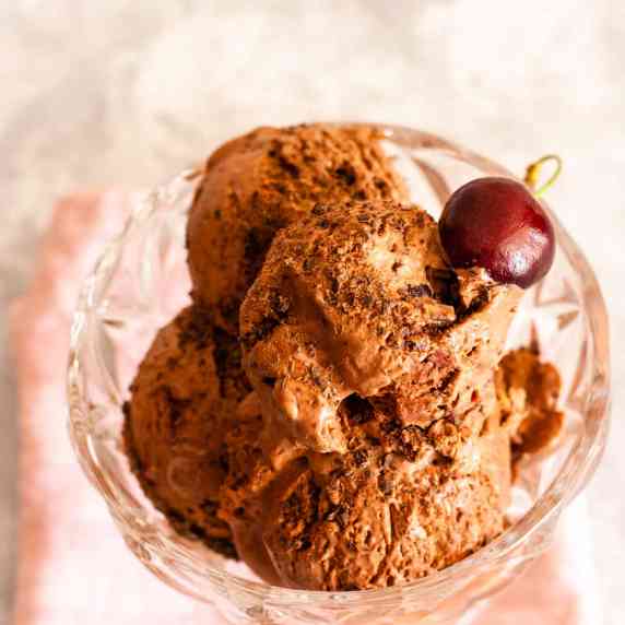 Scoops of black forest ice cream in a glass bowl topped with a cherry.