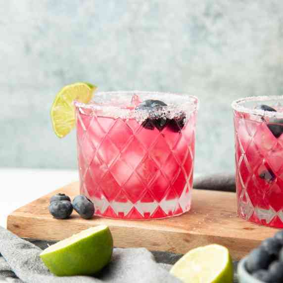 Two blueberry margaritas served in rocks glasses with salt rims, blueberries, and lime wedges.