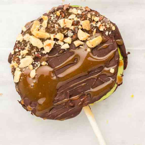 A slice of apple laying on parchment paper on a stick covered in caramel, chocolate and peanuts.