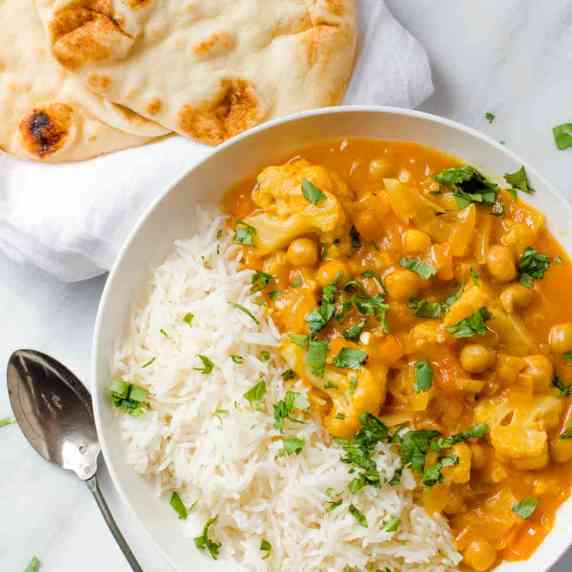 A bowl with white rice and chickpea and cauliflower curry stands with naan bread and a spoon nearby.