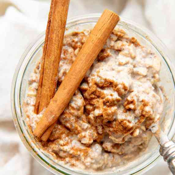 A spoon digs into a jar of chai latte overnight oats topped with cinnamon and two cinnamon sticks.