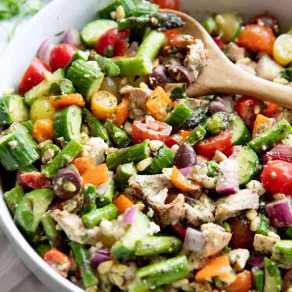 A wooden spoon scoops into a bowl of colorful chopped greek salad with grilled chicken and asparagus