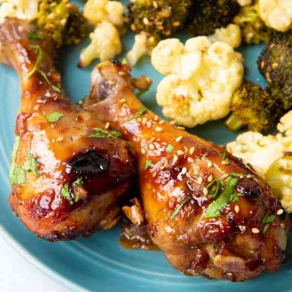 Close-up of two baked chicken drumsticks on a blue plate with roasted broccoli and cauliflower.