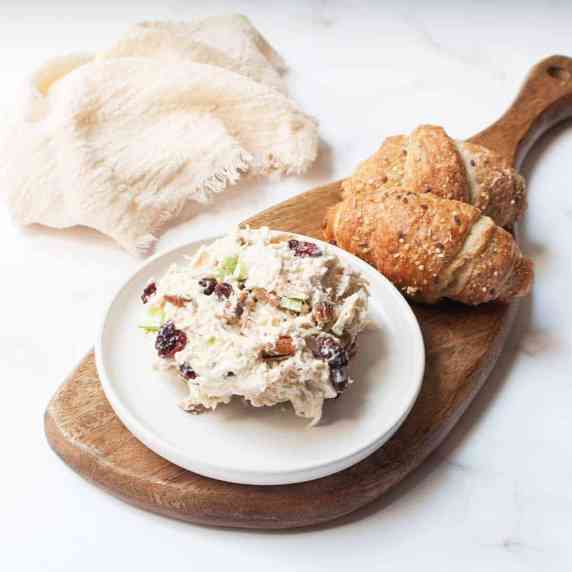 Chicken salad with cranberries and pecans on a white plate beside croissants on a wooden tray.