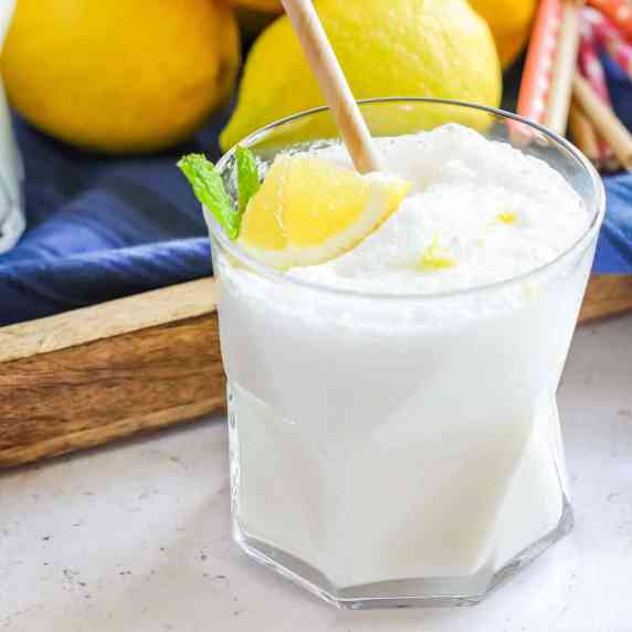 Close up side shot of frosted lemonade in a clear glass with two straws and a lemon and mint as a ga