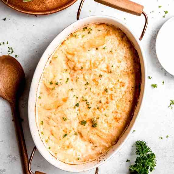 Creamy scalloped potatoes in a white baking dish on a light counter with a wooden spoon alongside.