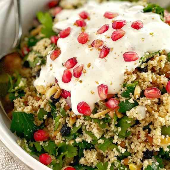 couscous salad garnished with yoghurt and pomegranate seeds in a white bowl with a spoon.