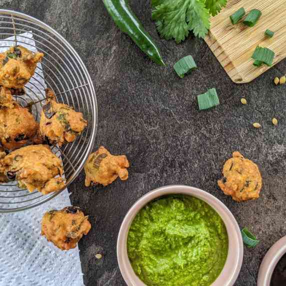 pakoras on a spider strainer on a grey background, green & tamarind chutney in a pink bowl