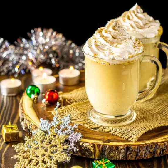 An eggnog latte with Christmas decor around it on a wooden platter.