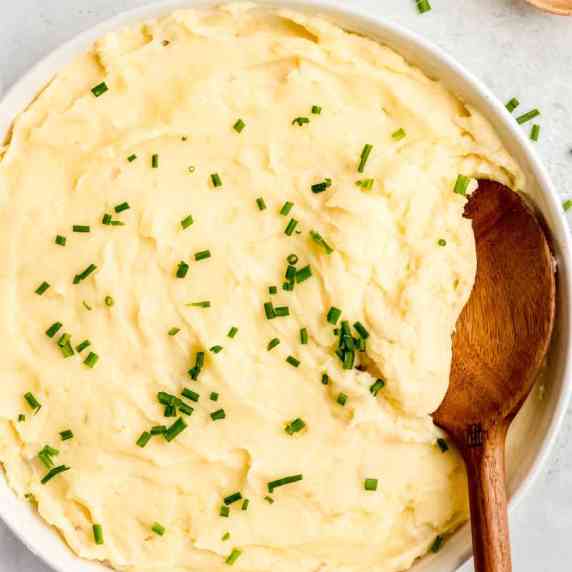 A spoon dips into creamy Instant Pot mashed potatoes served family-style in a large white bowl.