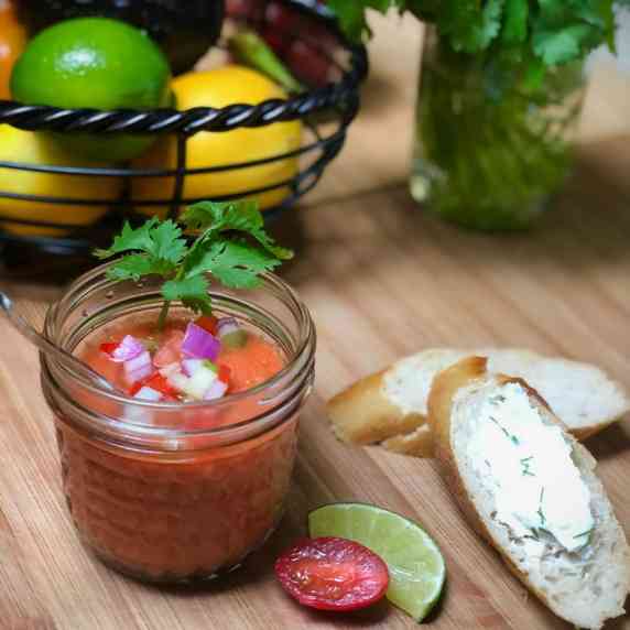 A glass jar filled with chilled gazpacho topped with fresh veggies and a side of garlic toast.