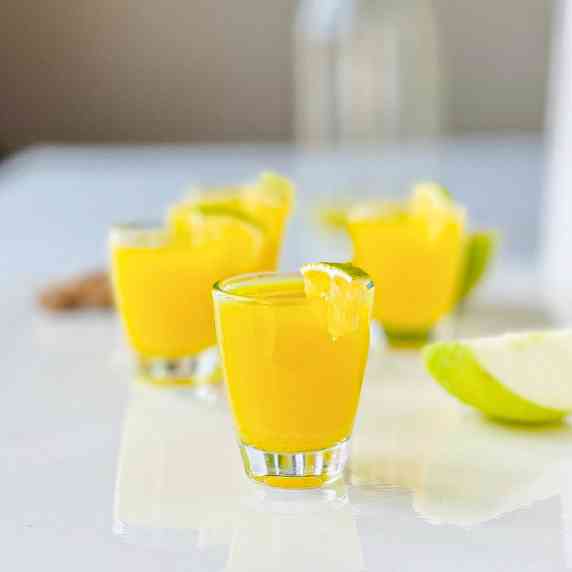 Ginger turmeric shots in shot glasses with orange wedges and a quartered apple in the background