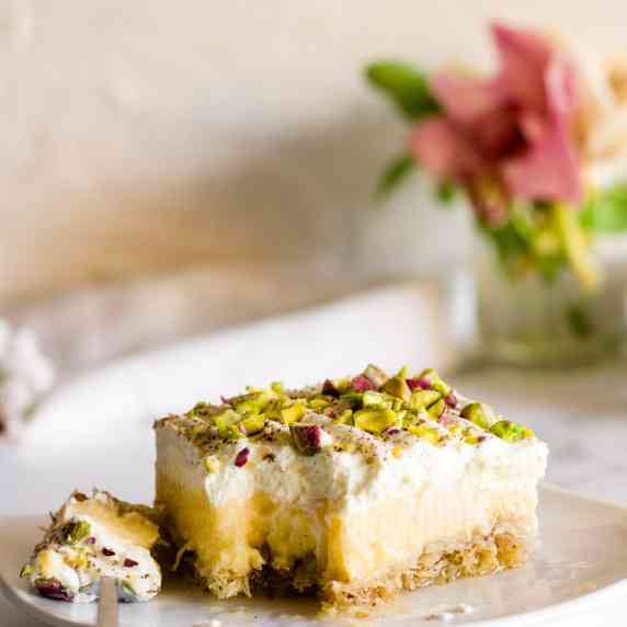 A slice of Greek ekmek kataifi dessert on a white plate with flowers in the background.
