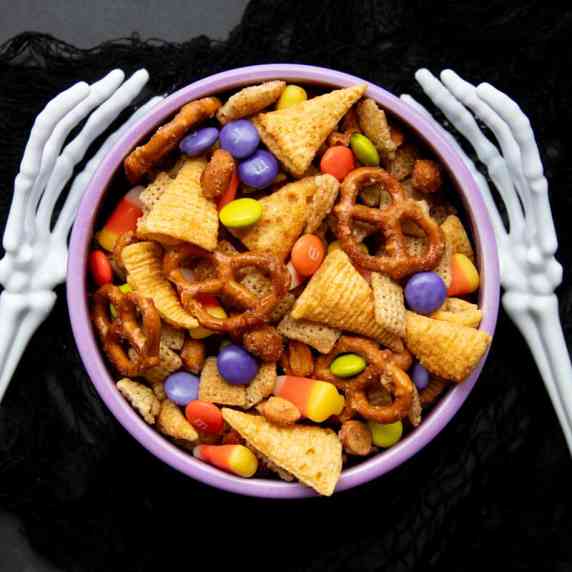 Two skeleton arms reach out, bony fingers curving around a bowl of Halloween Chex mix.