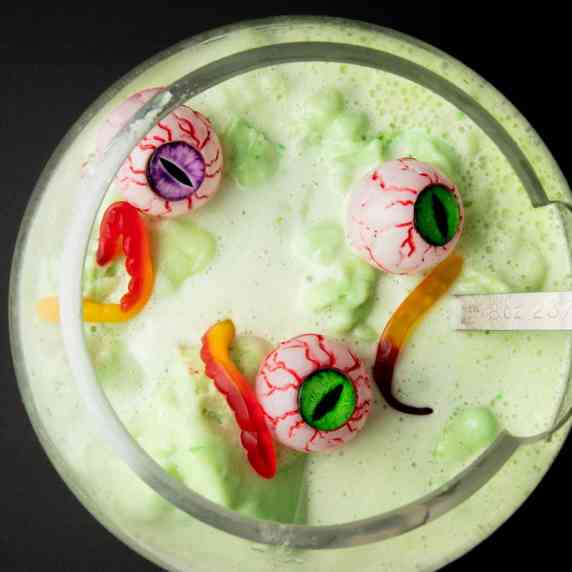 A large punch bowl filled with green Halloween punch garnished with gummy worms and bloodshot eyes.