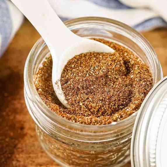Side close up shot of a white spoon scooping some chili seasoning out of a clear mason jar.