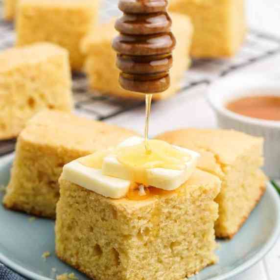 A honey dipper drizzles honey over a tall square of cornbread topped with two pats of butter.