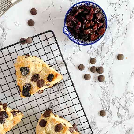 chocolate chip cherry scones sit on a cooling rack on a marble surface with a bowl of dried cherries